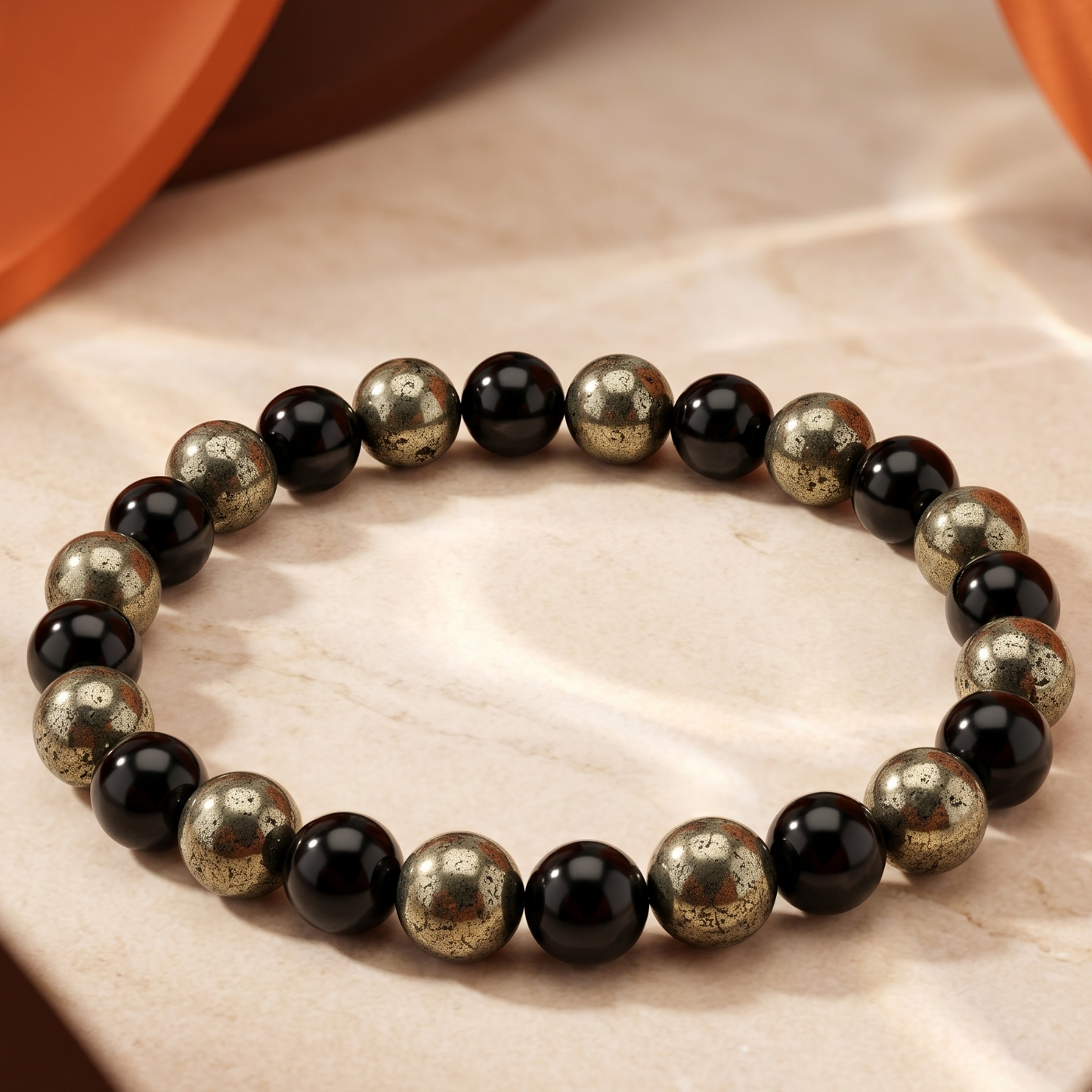A beaded stretch bracelet made of alternating polished black and metallic gold-toned round beads, displayed on a beige stone surface with warm orange-brown draped props in the background.