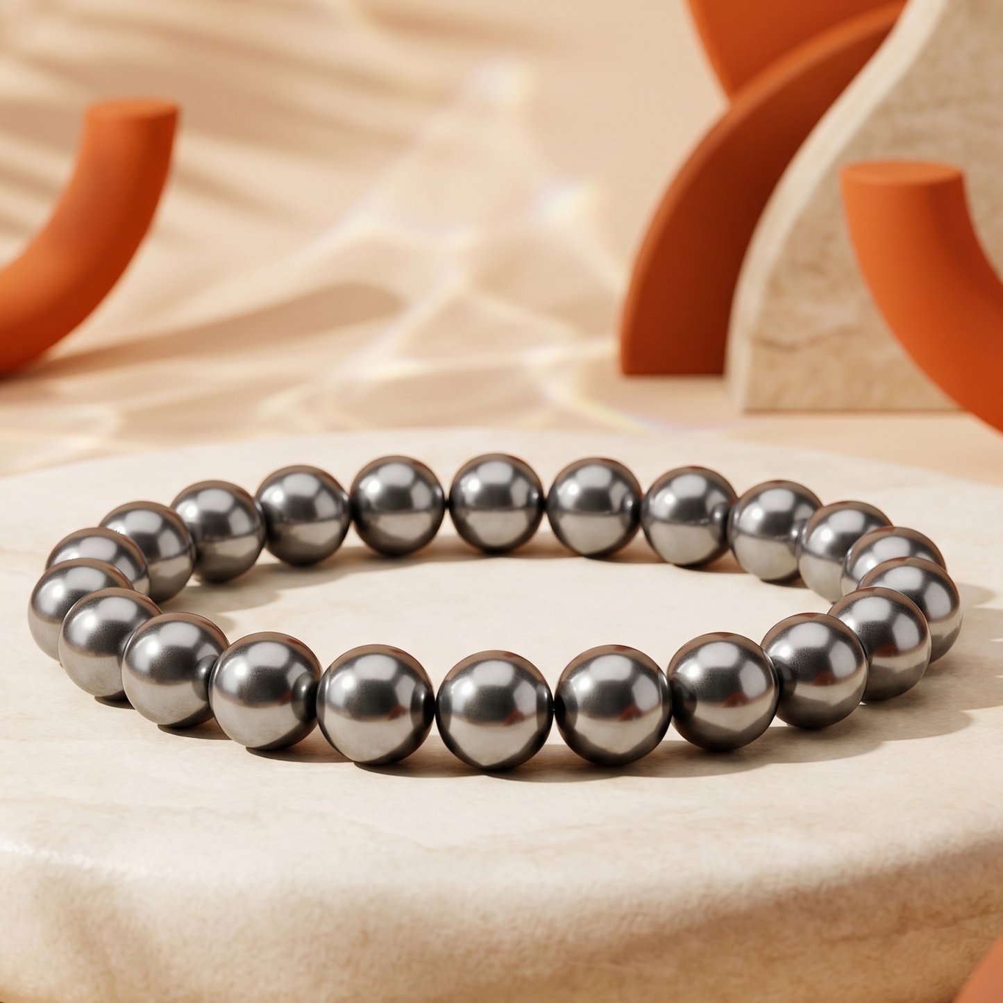 A beaded bracelet made of dark gray metallic beads, displayed on a round beige stone pedestal with warm, sunlit background props.