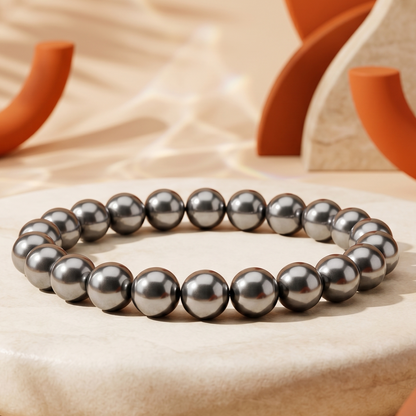 A beaded bracelet made of dark gray metallic beads, displayed on a round beige stone pedestal with warm, sunlit background props.