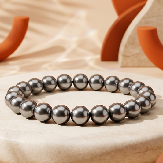A beaded bracelet made of dark gray metallic beads, displayed on a round beige stone pedestal with warm, sunlit background props.